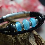 Memorial bracelet with two black braided leather bands and a light blue glass bead containing visible cremation ashes.