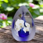 Design urn made of glass with transparent blue ash chamber and image of a white orchid – suitable for storing cremation ashes.