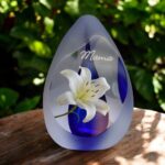 Design urn made of glass with transparent blue ash chamber and image of a white lily – suitable for storing cremation ashes.