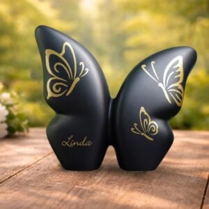 Black ceramic butterfly urn on wooden table in a forest, with gold foil butterflies and space for a name, suitable for cremation ashes.